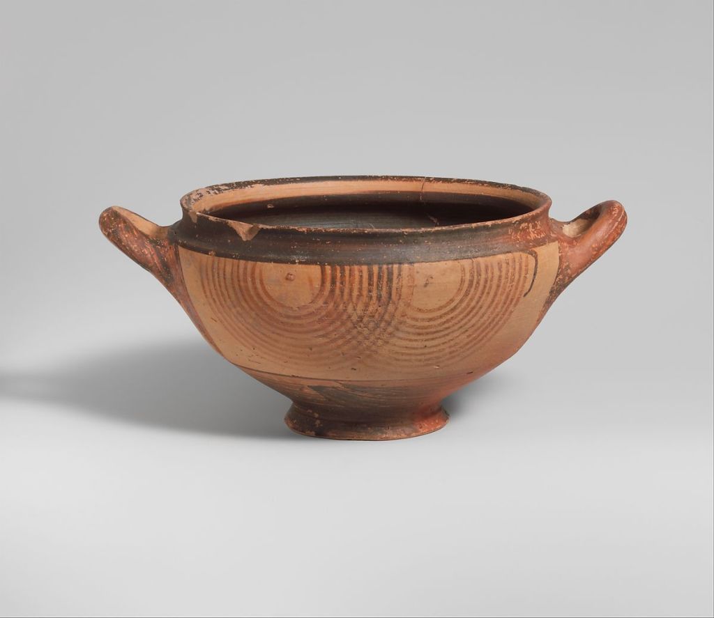 a clay skyphos drinking vessel with two handles and a wide mouth sits on a white table with a white background. It is various shades of brown and is decorated with concentric circles