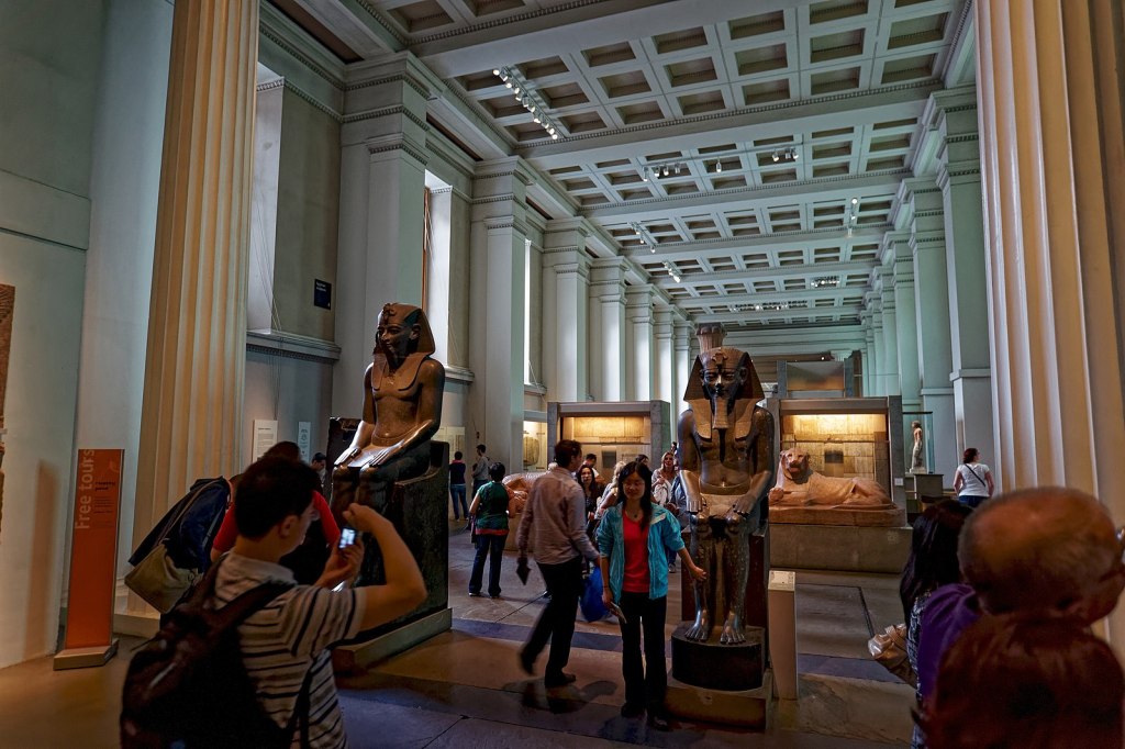 Large hallway of museum lined by pilasters on either side; Egyptian freestanding sculptures throughout along with glass display cases; people walking among these things; some taking pictures