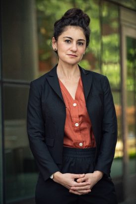 A professional photo of Dr. Anna Bonnell Freidin, a white woman which dark brown hair, which is piled on top of her head in a bun. She wears a black suit and an orange shirt. 