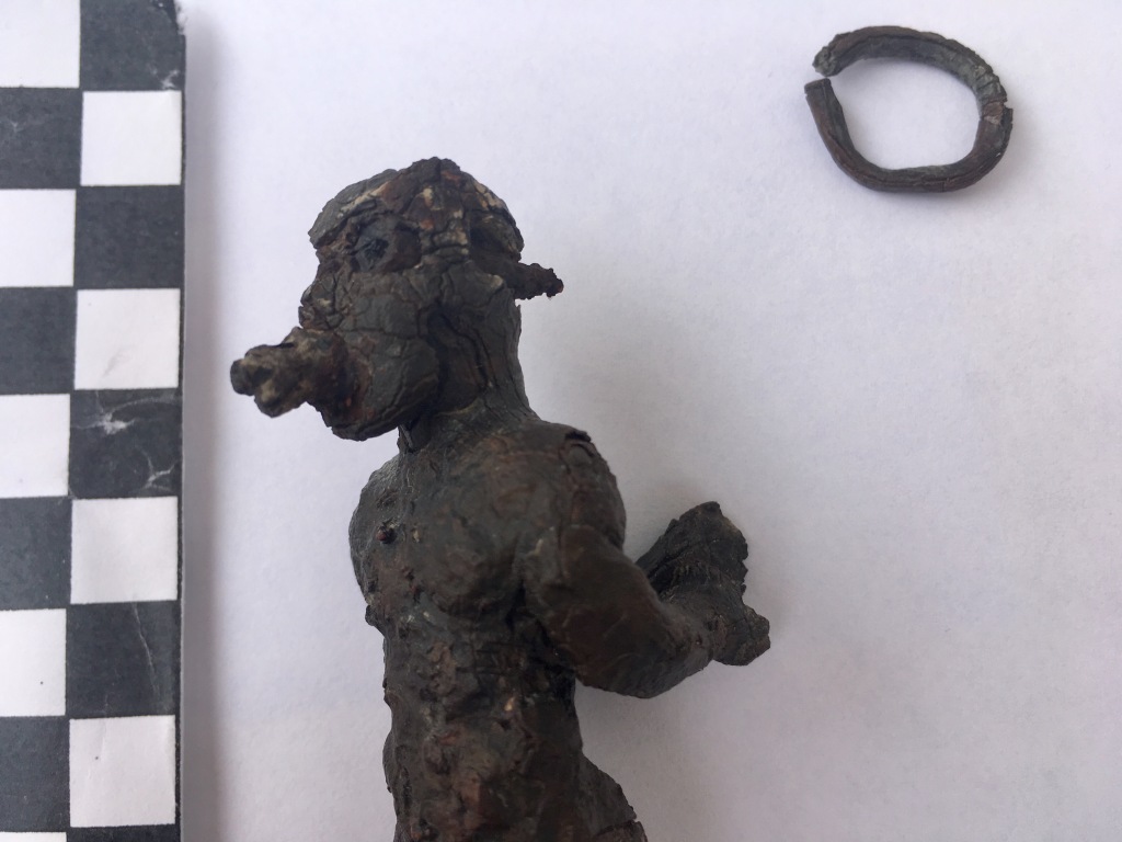 A lead effigy of a person with their arms bound behind their back. The photo also includes a small lead ring and a scale to measure the size of the objects. 