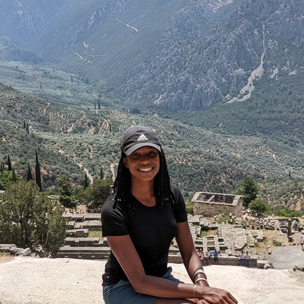 Image of Thelma Beth Minney sitting in front of the remains of the Temple of Apollo at Delphi. She is a Black woman, with long black hair, who wears a black shirt and black baseball hat. 