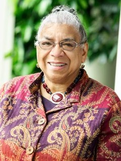 Head shot of Dr. Shelley Haley, a Black woman with grey hair and glasses, who wears a long-sleeved shirt with purple, pink and gold patterning. 